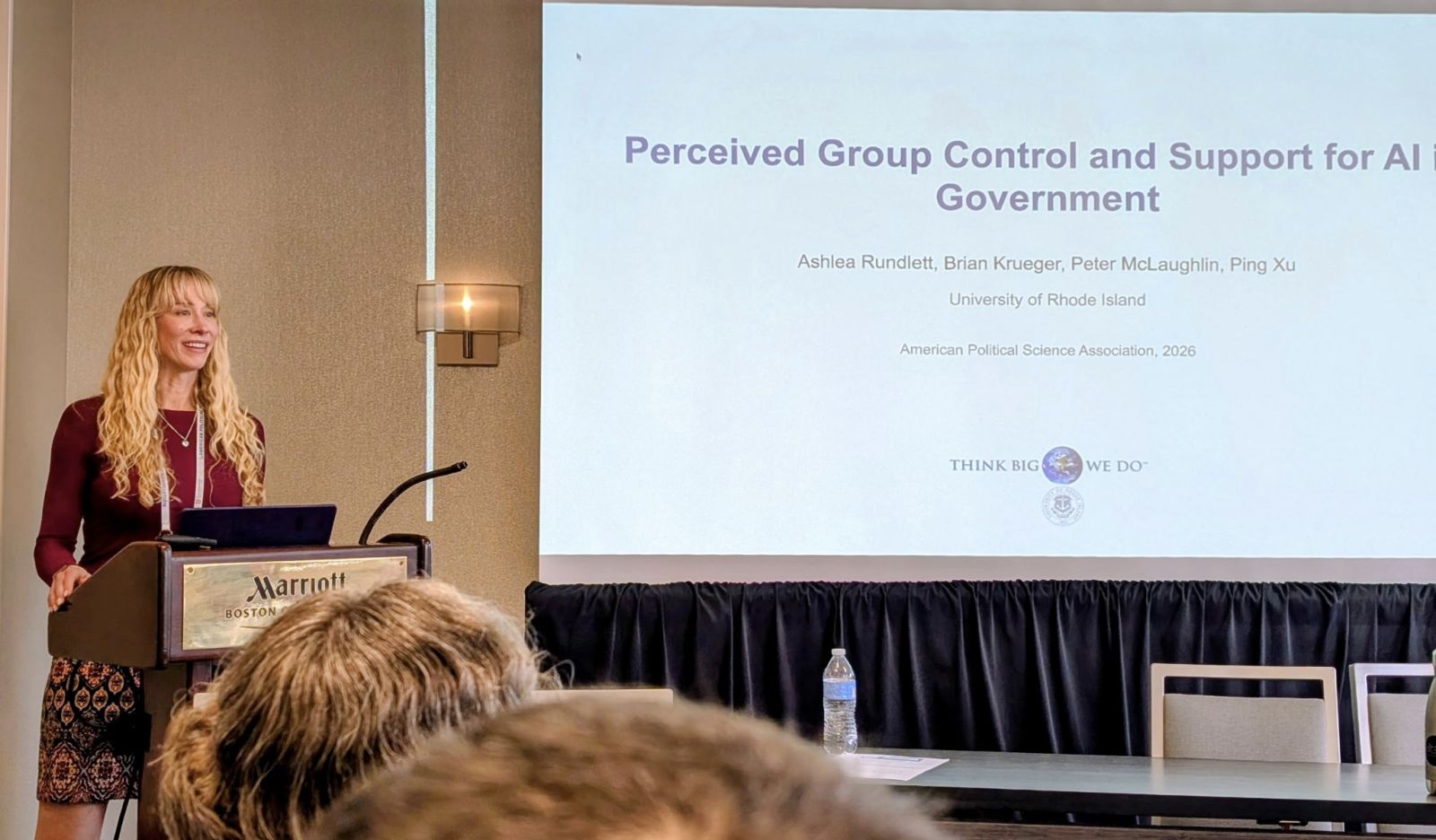 Ashlea Rundlett presenting at a podium at the 2026 American Political Science Association meeting in Boston, with a projected slide titled Perceived Group Control and Support for AI in Government.